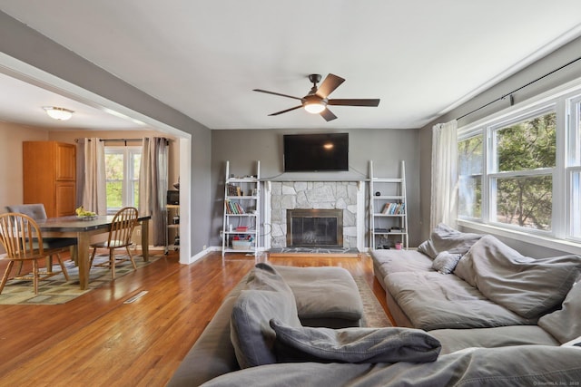 living area with wood finished floors, ceiling fan, a stone fireplace, and baseboards