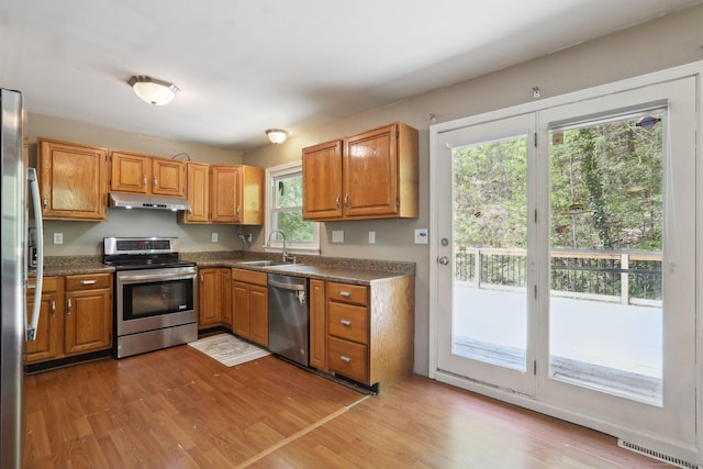 kitchen with appliances with stainless steel finishes, a sink, under cabinet range hood, dark countertops, and light wood-style flooring