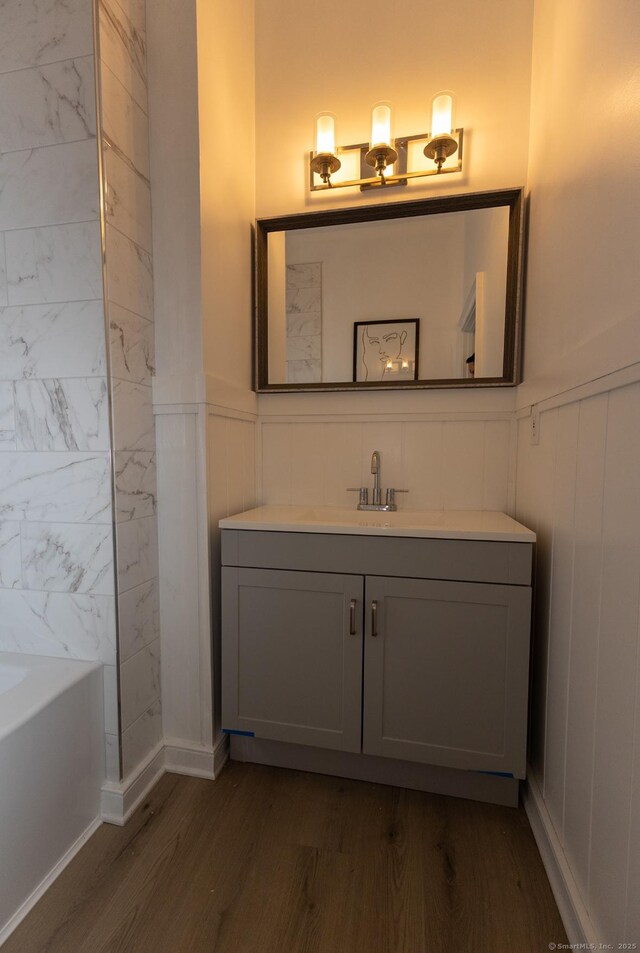 bathroom with wood finished floors, vanity, a bath, wainscoting, and a decorative wall