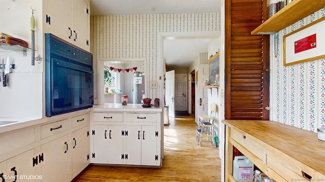 kitchen featuring wallpapered walls, light wood-type flooring, light countertops, white cabinets, and black oven