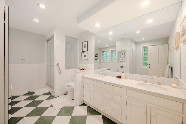 bathroom featuring a stall shower, toilet, wainscoting, double vanity, and tile walls