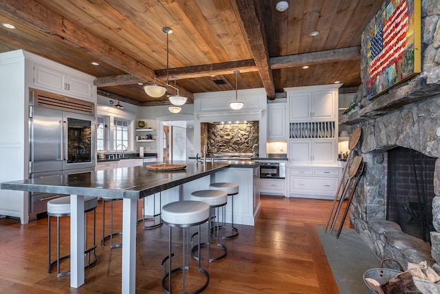 kitchen with dark countertops, wood ceiling with exposed beams, a stone fireplace, dark wood-style flooring, and stainless steel built in fridge