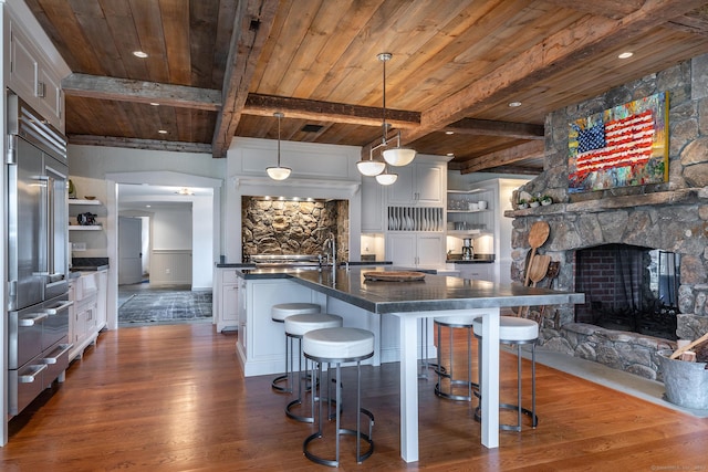 kitchen with stainless steel built in refrigerator, open shelves, dark wood finished floors, wood ceiling with exposed beams, and a stone fireplace