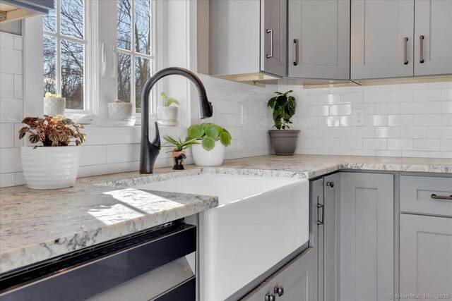 kitchen view of backsplash, gray cabinets, dishwasher, a sink, and light stone counters