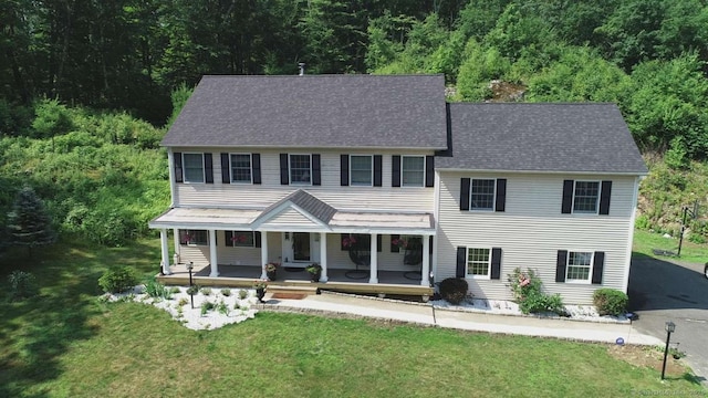 colonial-style house with a porch, a front yard, a view of trees, and roof with shingles