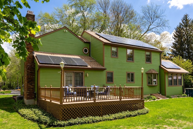 rear view of property featuring roof mounted solar panels, a deck, a yard, a chimney, and a shingled roof