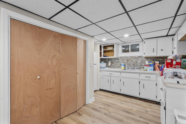 kitchen with a sink, light wood-type flooring, light countertops, a drop ceiling, and white cabinets