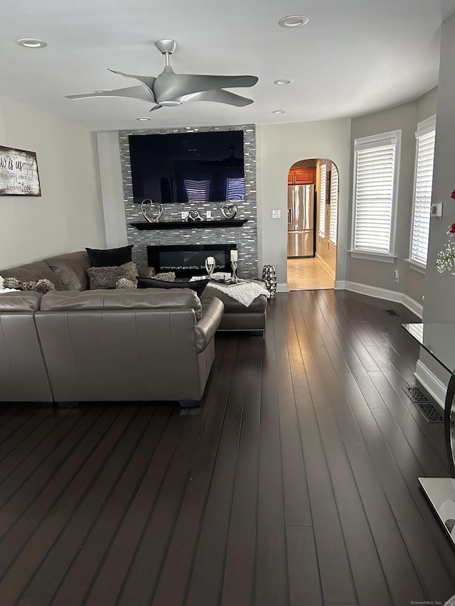 living area with a stone fireplace, ceiling fan, arched walkways, baseboards, and dark wood finished floors