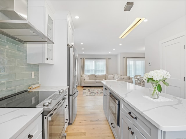 kitchen featuring stainless steel appliances, wall chimney exhaust hood, open floor plan, light wood-style floors, and glass insert cabinets