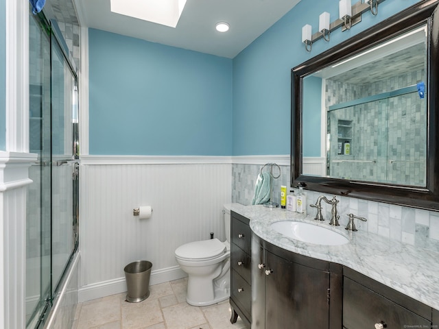 bathroom with a skylight, toilet, wainscoting, vanity, and bath / shower combo with glass door