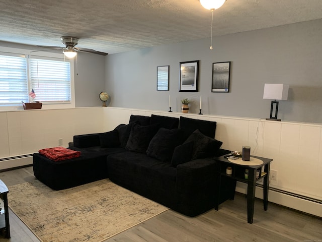 living area with a ceiling fan, a textured ceiling, wood finished floors, baseboard heating, and wainscoting