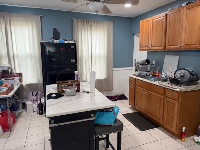 kitchen with a ceiling fan, freestanding refrigerator, light tile patterned flooring, a wainscoted wall, and light countertops