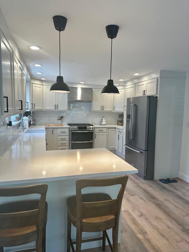 kitchen with stainless steel appliances, wall chimney range hood, a peninsula, light wood-type flooring, and light countertops