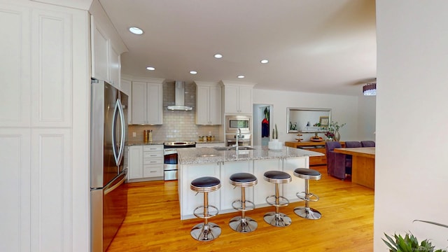 kitchen featuring appliances with stainless steel finishes, wall chimney range hood, light wood finished floors, a sink, and tasteful backsplash