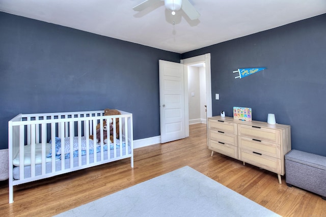 bedroom with wood finished floors, ceiling fan, baseboards, and a nursery area