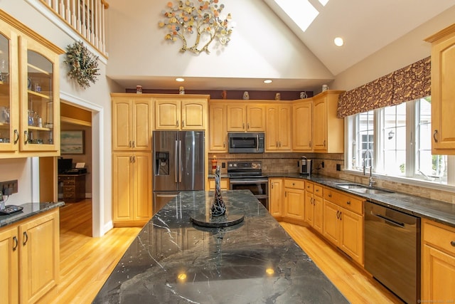 kitchen with appliances with stainless steel finishes, a sink, a skylight, light wood-type flooring, and dark stone countertops