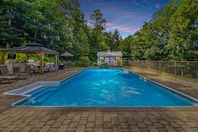 pool at dusk featuring a gazebo, a patio area, fence, and an outbuilding