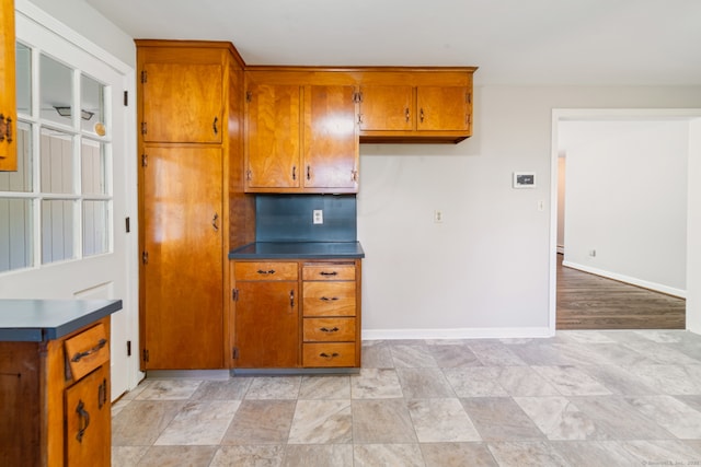 kitchen featuring brown cabinetry, dark countertops, baseboards, and light stone finish flooring