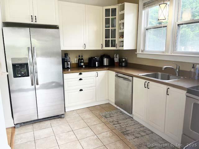 kitchen with stainless steel appliances, a sink, white cabinetry, glass insert cabinets, and light tile patterned floors
