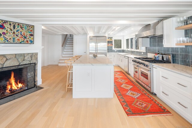 kitchen featuring premium appliances, wall chimney range hood, open shelves, light wood-type flooring, and backsplash