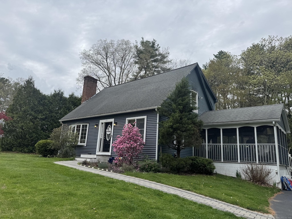 cape cod-style house featuring a sunroom, a chimney, a front yard, and a shingled roof