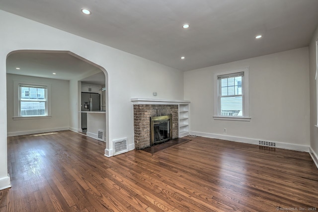 unfurnished living room with dark wood finished floors, a brick fireplace, arched walkways, baseboards, and recessed lighting