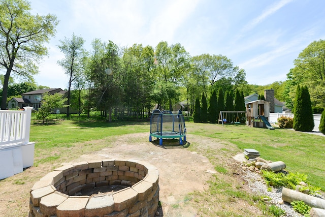 view of green lawn with a trampoline, a playground, and a fire pit