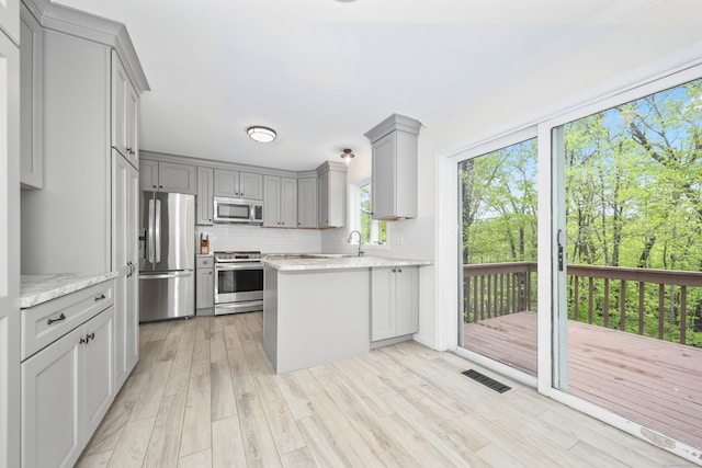 kitchen with appliances with stainless steel finishes, gray cabinets, a peninsula, light wood-style flooring, and tasteful backsplash