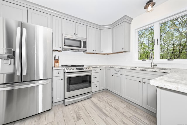 kitchen with appliances with stainless steel finishes, a sink, gray cabinetry, light stone countertops, and light wood finished floors