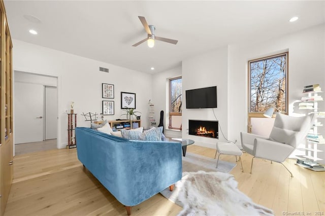 living area featuring light wood-type flooring, a ceiling fan, a warm lit fireplace, and recessed lighting