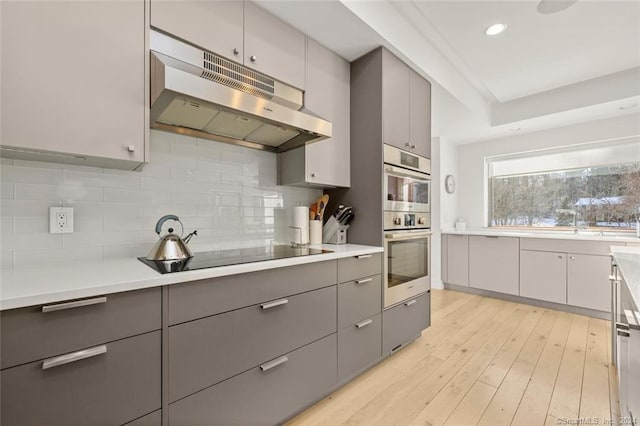 kitchen featuring under cabinet range hood, gray cabinetry, black electric stovetop, light wood finished floors, and backsplash