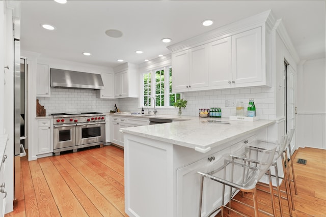 26 Heritage Ln, Weston, CT 06883 - kitchen featuring range with two ovens, wall chimney range hood, a peninsula, white cabinetry, and light wood finished floors
