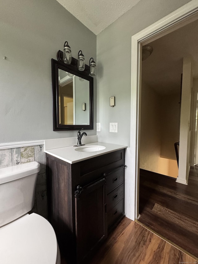 half bath featuring wood finished floors, toilet, vanity, a textured ceiling, and a wainscoted wall