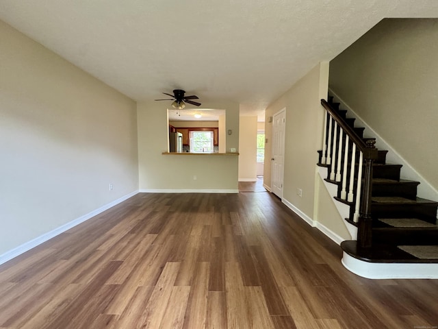 unfurnished living room with dark wood-style floors, stairs, a ceiling fan, and baseboards