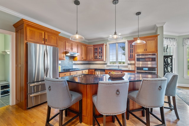 kitchen with stainless steel appliances, under cabinet range hood, brown cabinetry, backsplash, and glass insert cabinets
