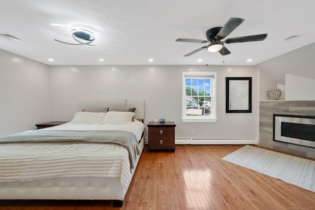 bedroom featuring baseboard heating, hardwood / wood-style floors, a fireplace, a ceiling fan, and baseboards