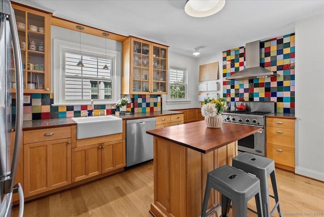 kitchen featuring appliances with stainless steel finishes, wall chimney range hood, a sink, butcher block countertops, and light wood finished floors