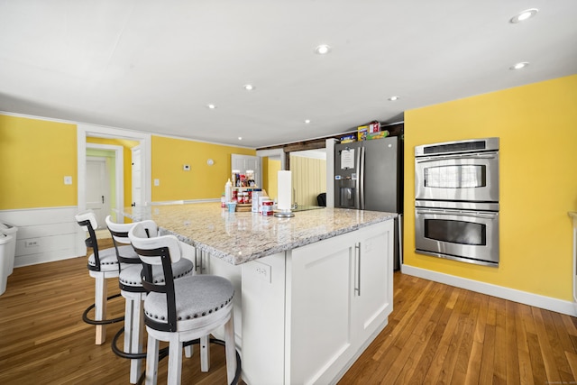 kitchen featuring stainless steel double oven, white cabinetry, a center island with sink, light stone countertops, and recessed lighting