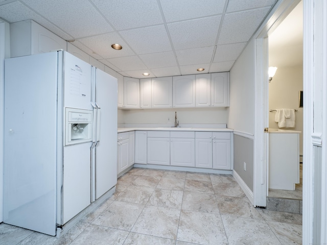 kitchen with white refrigerator with ice dispenser, a sink, a drop ceiling, light countertops, and white cabinets