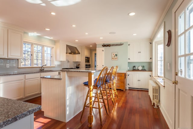 163 Lake Rd, Warren, CT 06754 - kitchen with radiator heating unit, wall chimney range hood, dark wood-style flooring, a breakfast bar area, and white cabinetry
