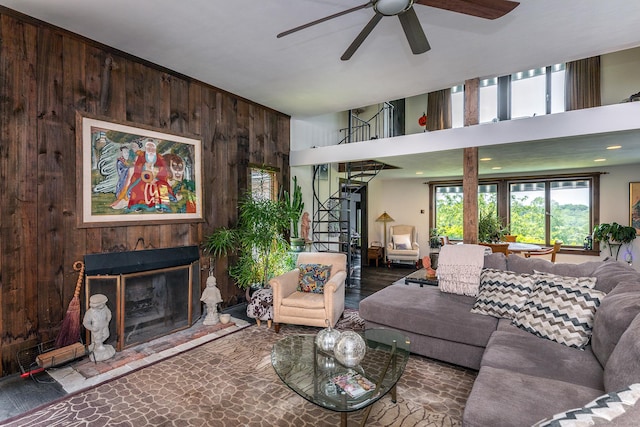 living area featuring wooden walls, ceiling fan, a fireplace with raised hearth, stairway, and a towering ceiling
