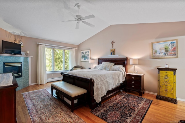 bedroom with a baseboard heating unit, lofted ceiling, light wood finished floors, a tiled fireplace, and ceiling fan
