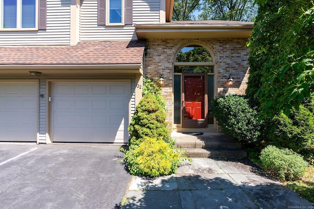 entrance to property featuring an attached garage, brick siding, asphalt driveway, and roof with shingles