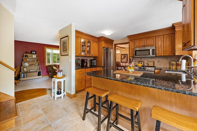 kitchen with stainless steel appliances, a peninsula, brown cabinetry, tasteful backsplash, and dark stone countertops