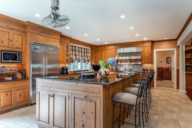 kitchen featuring built in appliances, wall chimney exhaust hood, ornate columns, a kitchen island, and crown molding
