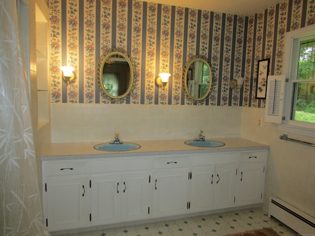 full bathroom featuring a baseboard radiator, wallpapered walls, tile walls, double vanity, and wainscoting