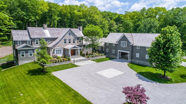 shingle-style home with an attached garage, stone siding, driveway, a front yard, and a chimney