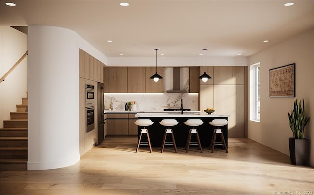 kitchen with pendant lighting, light wood-type flooring, wall chimney range hood, an island with sink, and a kitchen breakfast bar