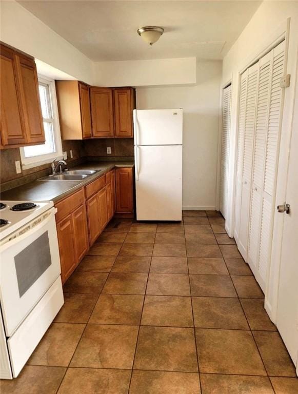 kitchen with white appliances, a sink, tile patterned floors, brown cabinetry, and dark countertops