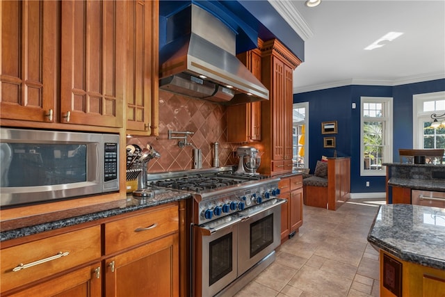 kitchen with wall chimney range hood, dark stone countertops, ornamental molding, appliances with stainless steel finishes, and tasteful backsplash